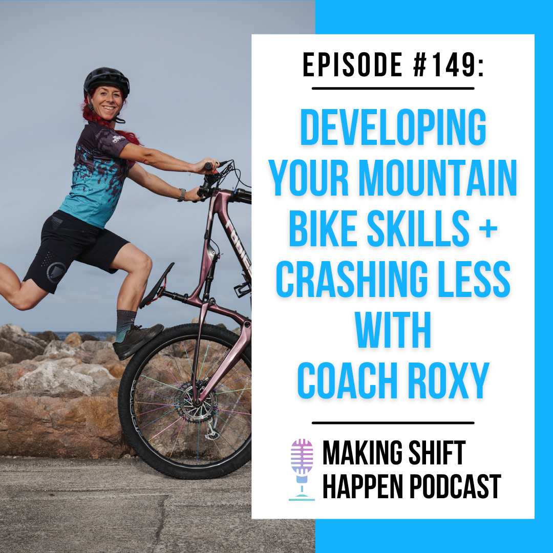 Coach Roxy is wearing black shorts and a black and teal jersey with a black helmet and red hair as she stands on top of the front wheel of her mountain bike, with a big smile across her face.