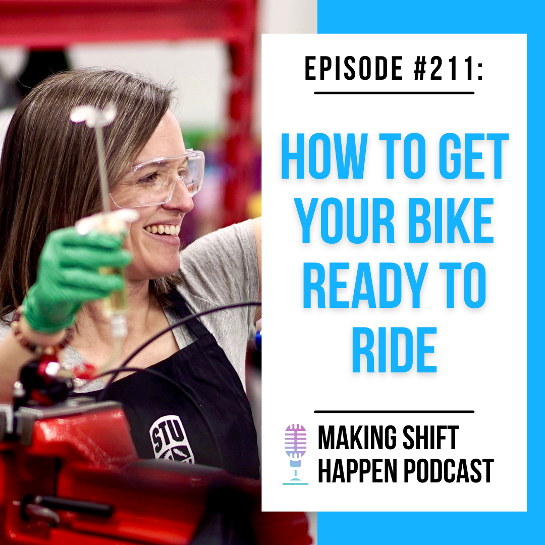 Jen is wearing safety glasses and a black apron while she is bleeding her bike's brakes with a smile on her face.